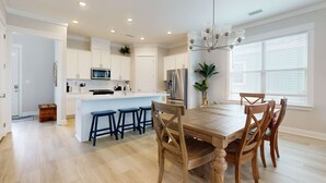A modern kitchen with white cabinets, a central island, bar stools, and a dining table with wooden chairs. - Brand-new home near the beach with on-site pool, central AC, & washer,dryer (Orange Beach)