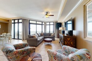 A spacious living room with a ceiling fan, a sectional sofa, patterned armchairs, a coffee table, and a TV on a wooden cabinet. - Spacious condo with pool, hot tub, tennis courts, & sauna (Orange Beach)