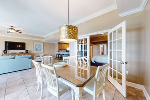 A spacious living room with a dining area, a television, and a kitchen in the background. - Spacious condo with pool, hot tub, tennis courts, & sauna (Orange Beach)