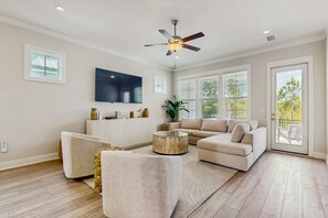 A spacious living room with a large flat-screen TV, a ceiling fan, and a sliding glass door leading to an outdoor area. - Modern beach house with porches, private washer and dryer- walk to the beach (Orange Beach)
