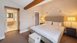 A bedroom with a large bed, wooden ceiling beams, a nightstand with a lamp, and a door leading to a bathroom.