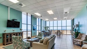 A spacious room with a large window, a flat-screen TV, and a dining area. - Renovated beachfront condo with balcony, central AC, pool, hot tub & tennis (Orange Beach)