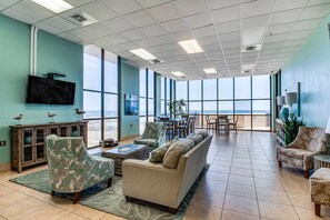 A spacious room with a large window, a flat-screen TV, and a dining area. - Renovated beachfront condo with balcony, central AC, pool, hot tub & tennis (Orange Beach)