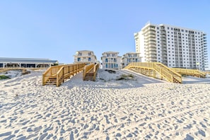 A beachside hotel with wooden boardwalks leading to the beach, a large building with multiple balconies, and a clear blue sky. - Spacious beachside community home with ocean view, private pool, & fireplace (Orange Beach)