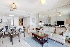 A modern living room with a sofa, coffee table, dining table, chairs, and a kitchen area with a refrigerator. - Beautiful Condo in Beachfront Resort w/WiFi, Central AC, Shared Pool & Hot Tub (Orange Beach)