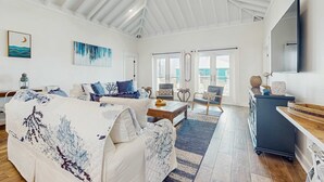 Ein geräumiges Wohnzimmer mit hoher Decke, einem großen Fenster, einer Sofagarnitur, einem Couchtisch und einem an der Wand montierten Fernseher.