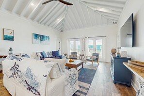 A spacious living room with a high ceiling, a large window, a sofa set, a coffee table, and a TV mounted on the wall. - Gulf-Front Oasis: Rooftop Heated Pool, Elevator & Dog-Friendly! (Dauphin Island)