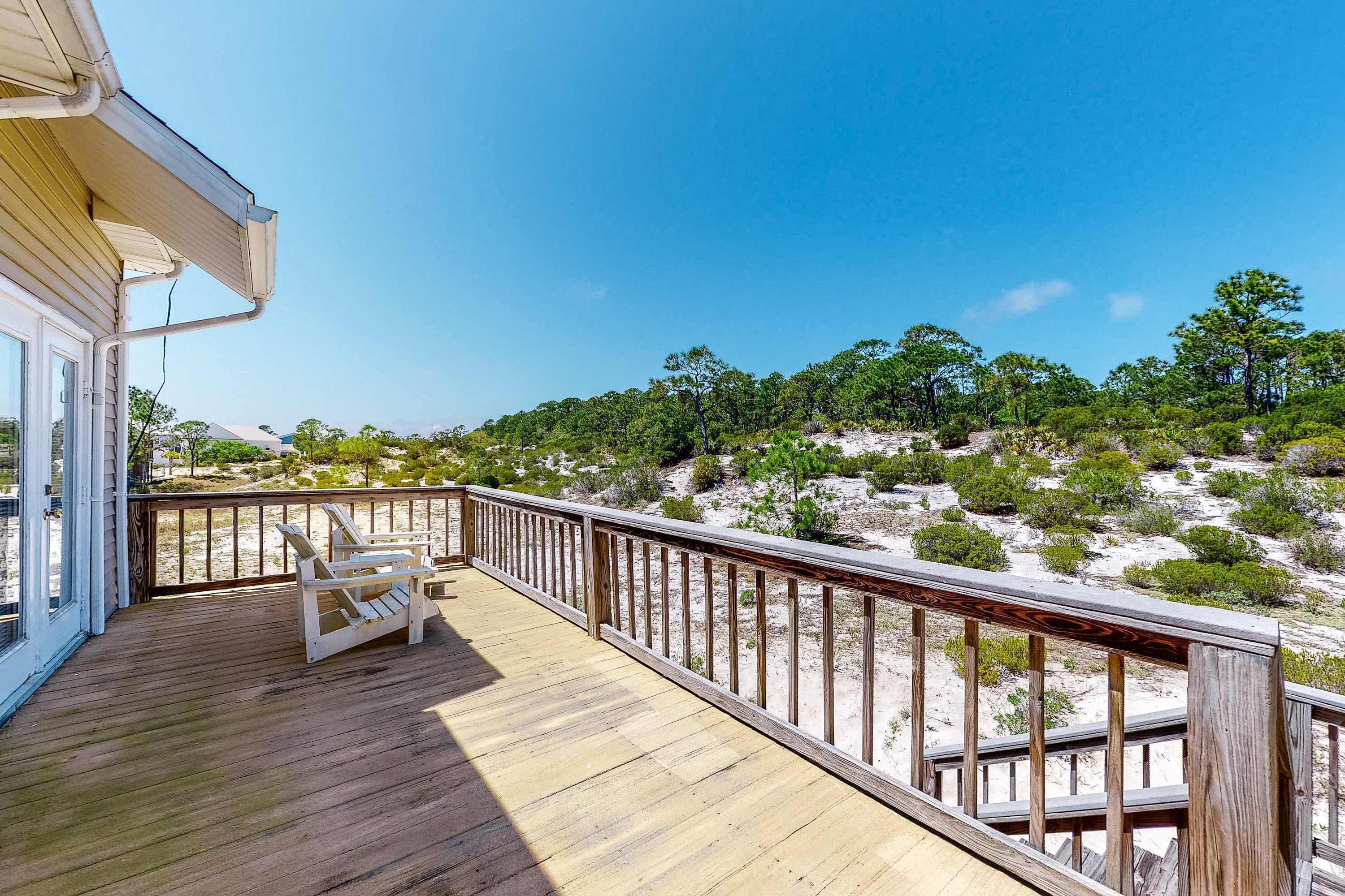 Eine Holzterrasse mit Geländer, ein weißer Stuhl und Blick auf einen sandigen Strand mit spärlicher Vegetation.