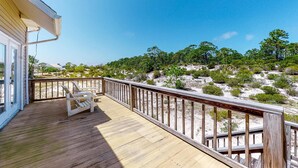 Eine Holzterrasse mit Geländer, ein weißer Stuhl und Blick auf einen sandigen Strand mit spärlicher Vegetation.