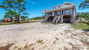 Casa de dos pisos frente a la playa con balcón, una escalera y una playa arenosa al frente.