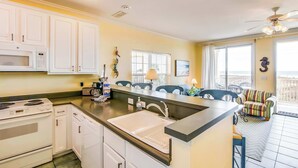A kitchen with white cabinets, a microwave, stove, and sink. There is a dining area with chairs and a view of the outside. - Gulf-front condo w/ lock-out feature, shared pools & amazing views (Dauphin Island)