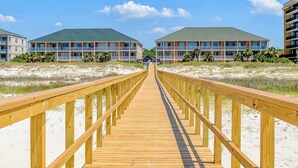 A wooden boardwalk leading to a beachfront hotel with multiple balconies and a clear sky. - Gulf-front condo w/ lock-out feature, shared pools & amazing views (Dauphin Island)