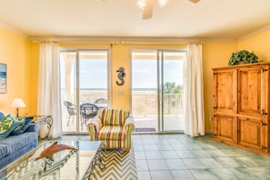Ein Wohnzimmer mit einer Couch, einem gestreiften Sessel, einem Glastisch, einem Holzschrank und Blick auf den Balkon.