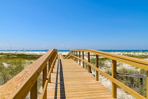Ein hölzerner Steg führt zu einem Strand mit Dünen und einem klaren, blauen Himmel.