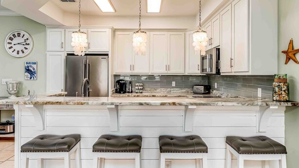 Una cocina con gabinetes blancos, un refrigerador de acero inoxidable, una encimera de mármol y tres taburetes de bar.