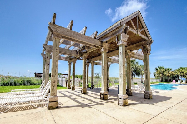 A wooden pergola structure with a pool and lounge chairs in the background.