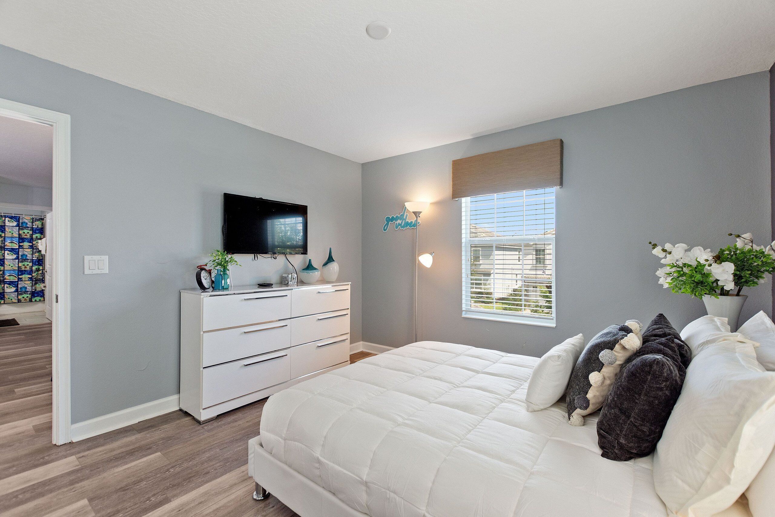 Un dormitorio con cama, cómoda, televisor, ventana con persianas y lámpara de pared.