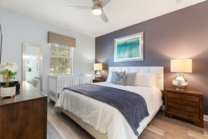 Une chambre à coucher avec un lit, des tables de chevet, un ventilateur de plafond, une fenêtre avec des stores et un tableau au mur.