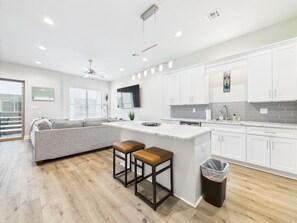 A modern kitchen with white cabinets, a grey island with stools, and a large window with blinds.