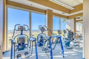 A gym with cardio machines, large windows, and a view of the beach. - Beachfront condo with stunning views, shared hot tub, fitness room, & indoor pool (Gulf Shores)