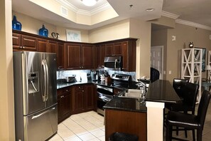 A kitchen with wooden cabinets, stainless steel appliances, a black countertop, and a dining area with a black table and chairs. - Gorgeous beachfront condo, shared pool and beach access (Gulf Shores)