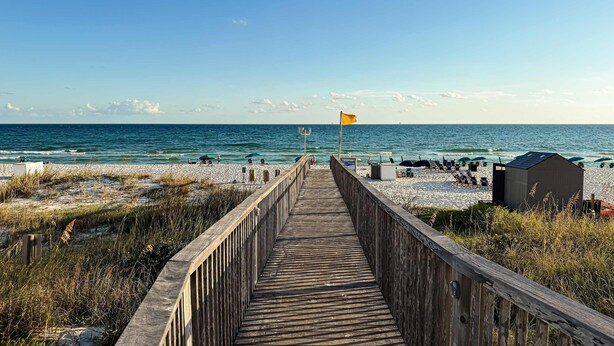 A wooden boardwalk leading to a beach with a flag, a lifeguard tower, and a small building. - Spacious beachfront condo with full kitchen, shared pool and beach access (Gulf Shores)