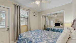 Een slaapkamer met een bed, een plafondventilator, een raam met zonwering en een spiegel die de kamer weerspiegelt.