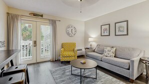 Een woonkamer met een bank, een gele fauteuil, een ronde salontafel, een klok en een trap zichtbaar door een glazen deur.