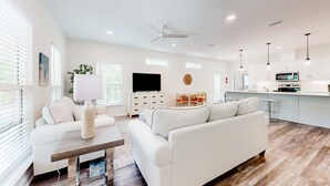 A spacious living room with a white sofa, a coffee table, and a TV. There is a kitchen area with a microwave and a bar.