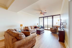 A spacious living room with a large sectional sofa, a coffee table, and a television. There is a ceiling fan, a window with a view, and a balcony. - Sunlit beach condo with pool, hot tub, & amazing view - steps to the sand (Dauphin Island)