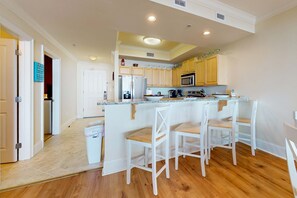 A modern kitchen with wooden cabinets, a central island with bar stools, and a built-in microwave. - Sunlit beach condo with pool, hot tub, & amazing view - steps to the sand (Dauphin Island)