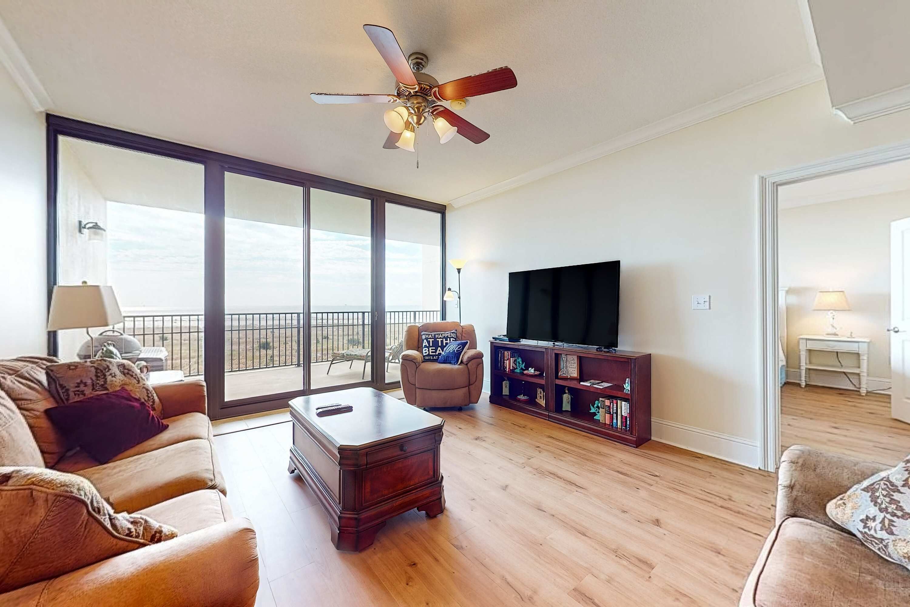 Een ruime woonkamer met een groot raam, een plafondventilator, een flatscreen-televisie, een boekenplank en een comfortabele bank.