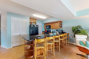 A kitchen with a central island, wooden chairs, a white chair, and a potted plant. - Beachfront 2-story condo with beach views, 2 pools, gazebo, balconies, W/D, & AC (Dauphin Island)