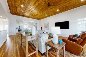 A spacious living area with a wooden ceiling, a kitchen in the background, and a large flat-screen TV mounted on the wall. - Inviting waterfront home with balcony, full kitchen, central AC, & W/D (Dauphin Island)