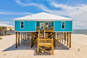 A blue beach house with a white roof, wooden deck, and a staircase leading to the entrance.