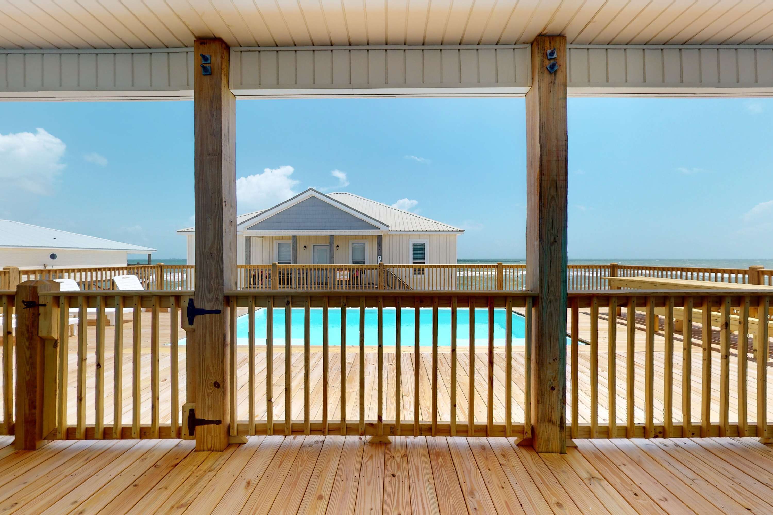 Una terraza de madera con piscina, una casa al fondo y cielo despejado.