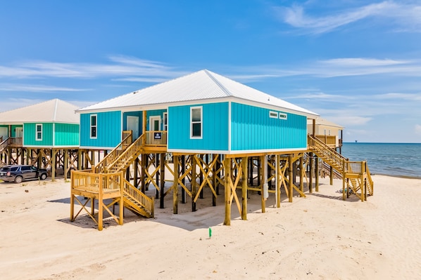 A beachfront wooden structure with a staircase leading to the entrance, a car parked nearby, and a clear sky.