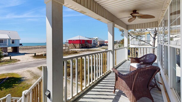 Eine überdachte Veranda mit Korbmöbeln, einem Deckenventilator und Blick auf den Strand und Häuser.