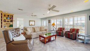 Ein Wohnzimmer mit Ledersessel, einer Couch, einem Couchtisch und zwei Sesseln. Es gibt Deckenventilatoren, ein Bücherregal und ein Fenster mit Blick.