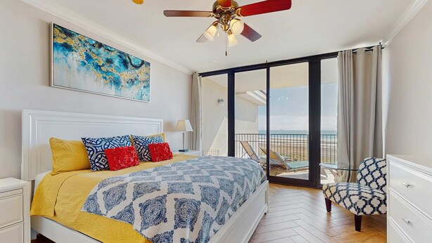 Une chambre avec un lit, un ventilateur de plafond, un tableau, un balcon avec une chaise et une vue sur l’extérieur.