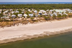 Luftaufnahme einer Küstenwohngegend mit Häusern, einem Strand und bewaldeter Landschaft im Hintergrund.