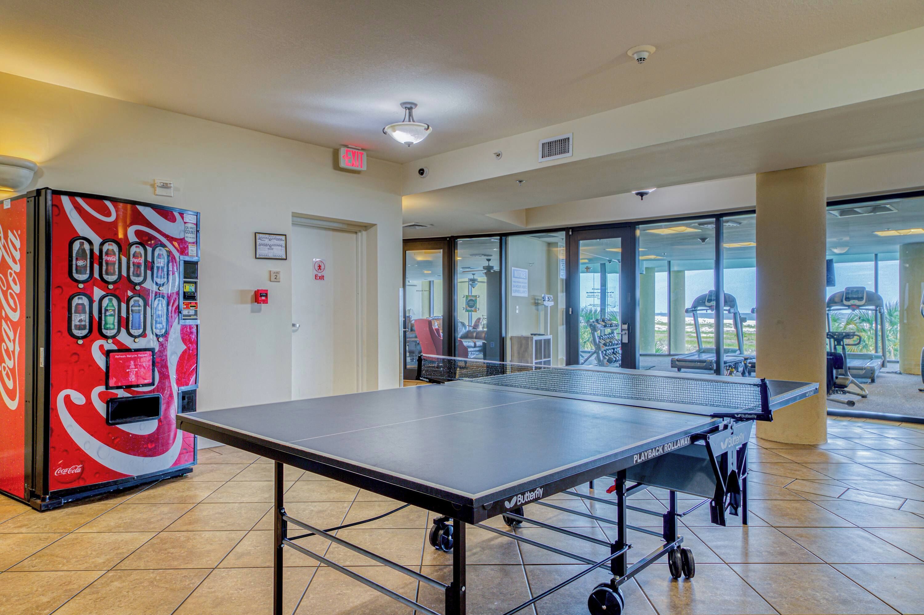 A ping pong table in a room with a Coca-Cola vending machine and a glass door leading to an outdoor area with exercise equipment.