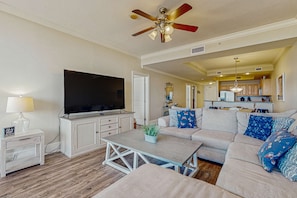 A living room with a TV, sofa, coffee table, and ceiling fan. - Lovely Gulf front condo w/shared sauna, hot tub, indoor pool, & stunning views (Dauphin Island)