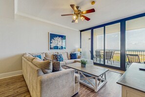 A modern living room with a sectional sofa, a coffee table, and a ceiling fan. - Lovely Gulf front condo w/shared sauna, hot tub, indoor pool, & stunning views (Dauphin Island)