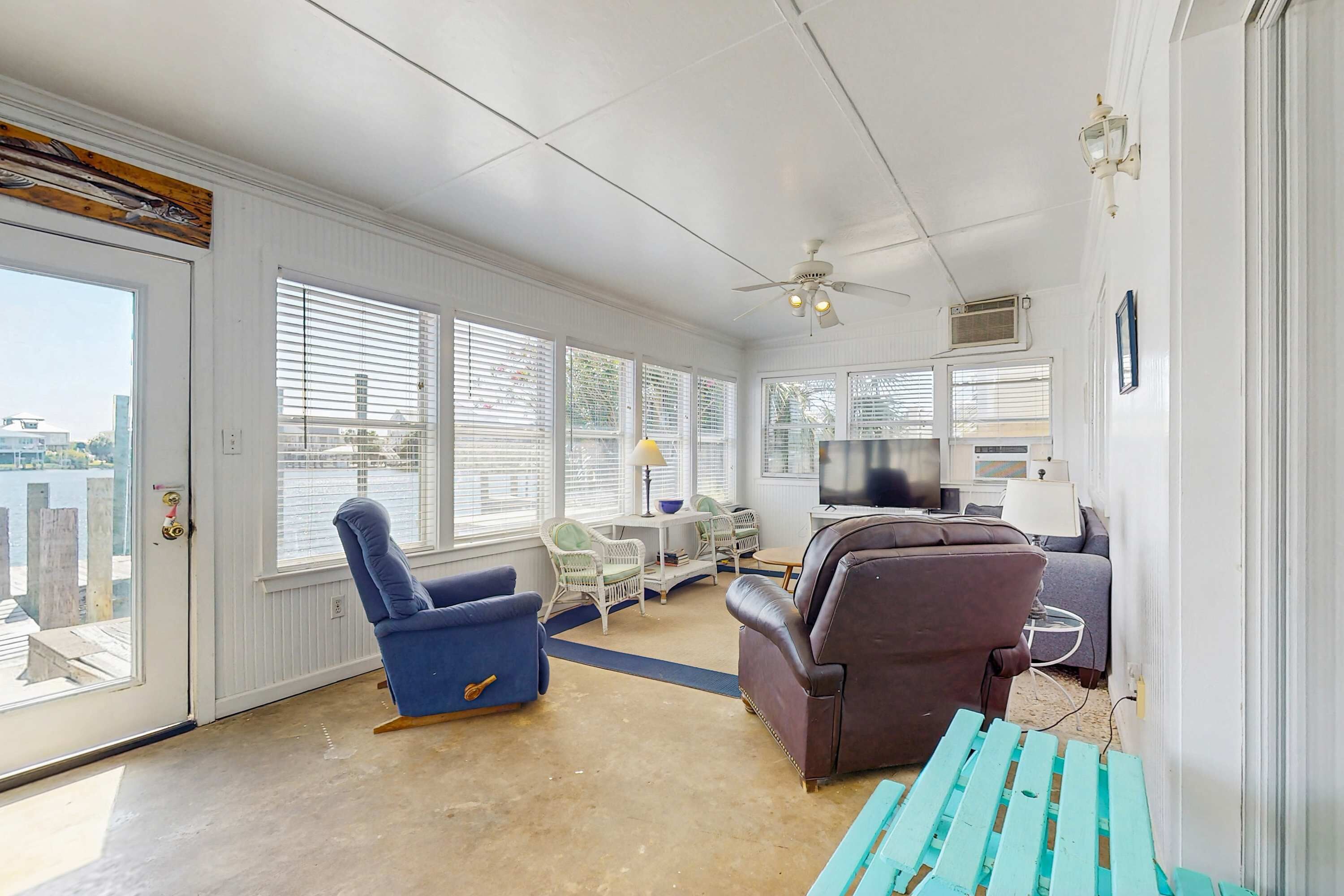 A spacious room with a ceiling fan, large windows with blinds, a blue armchair, a brown sofa, and a teal bench.