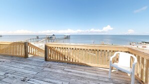 A wooden deck with a white plastic chair, overlooking a beach with a wooden pier and a clear blue sky. - Welcoming home with WiFi, gulf views & spacious deck - steps to the beach (Dauphin Island)