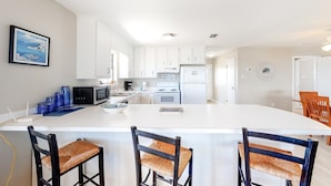 A modern kitchen with white cabinets, a central island, and a dining area with wooden chairs. - Welcoming home with WiFi, gulf views & spacious deck - steps to the beach (Dauphin Island)
