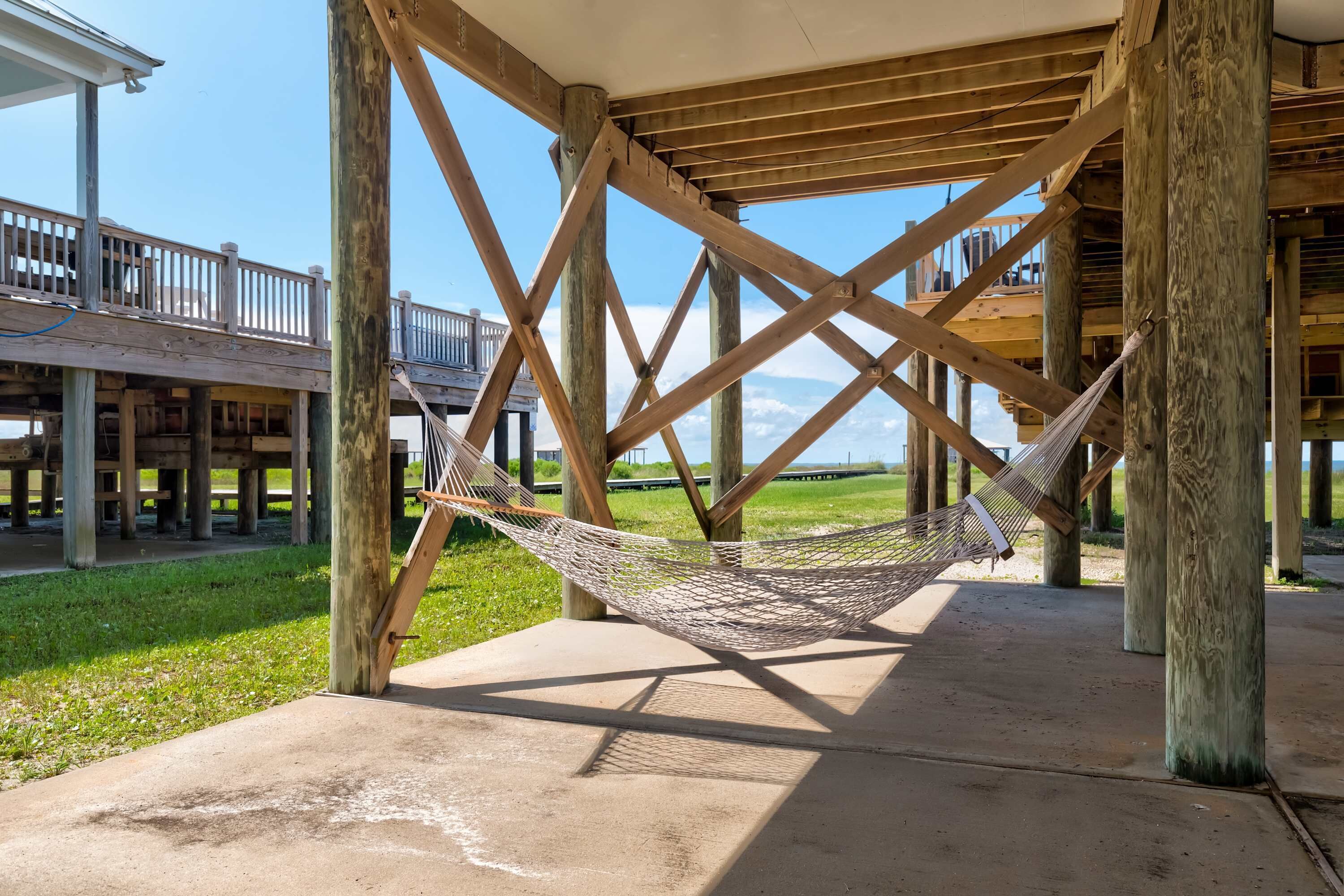 A hammock under a wooden structure with a view of a grassy area and a building in the background.