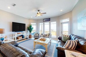 A living room with a leather sofa, a glass coffee table, a TV, and a ceiling fan. - Bayfront home with beach access & breathtaking water views - dog-friendly (Dauphin Island)