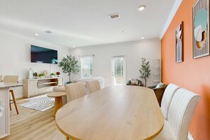 Ein modernes Wohnzimmer mit Esstisch aus Holz, weißen Sofas, einem Flachbildfernseher und einem großen Fenster.